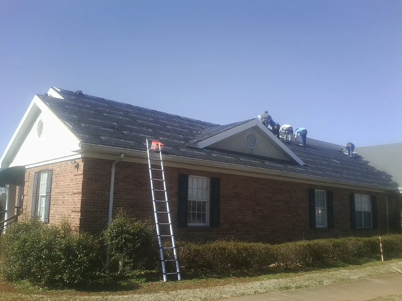 Crew tearing shingles off a brick ranch roof with a ladder set — Metal Roof Repair work by our crew in Braithwaite
