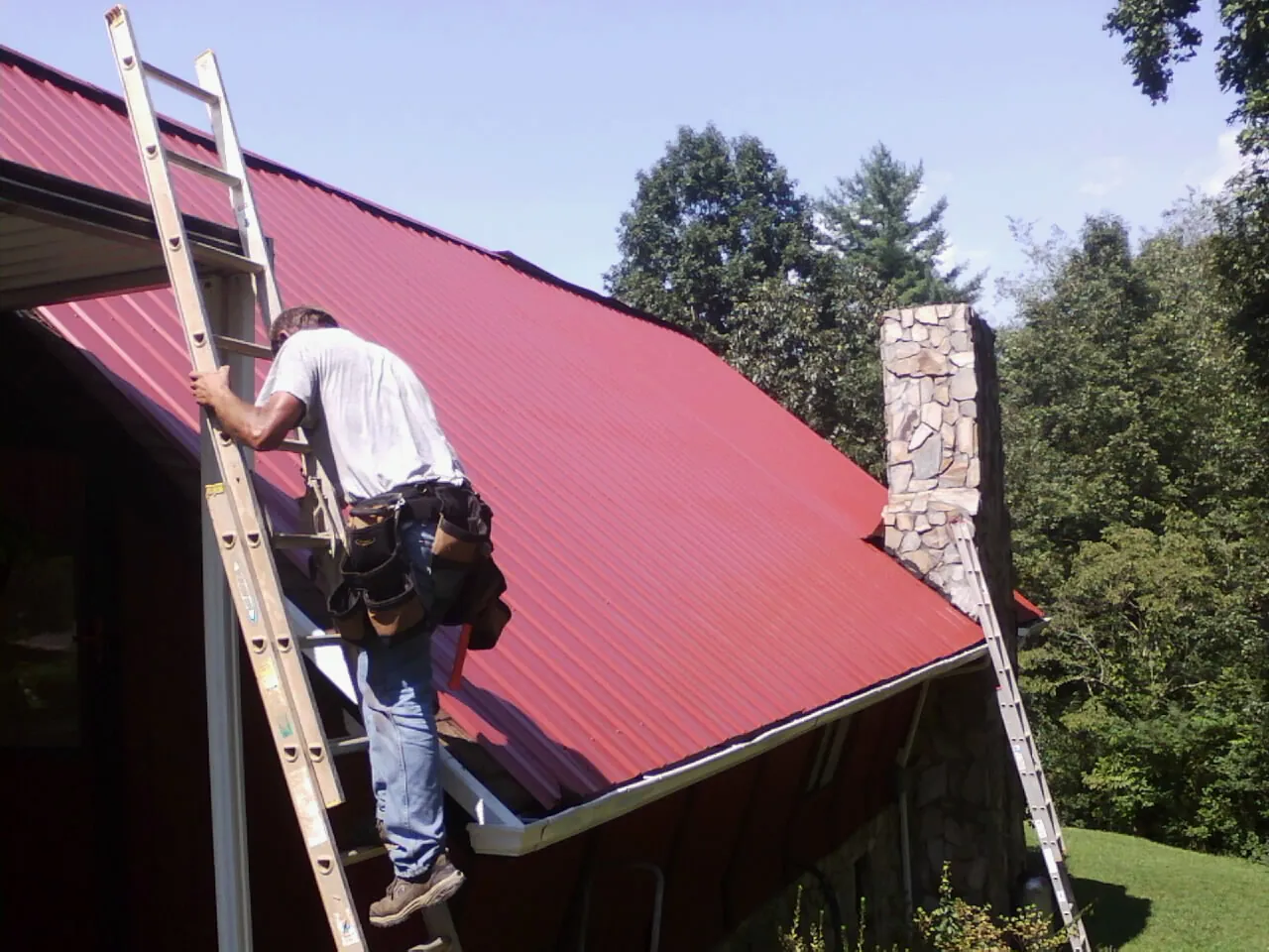Roofer on a ladder beside a red metal roof and stone chimney — Soffit & Fascia Repair by Roofing Brothers in Braithwaite